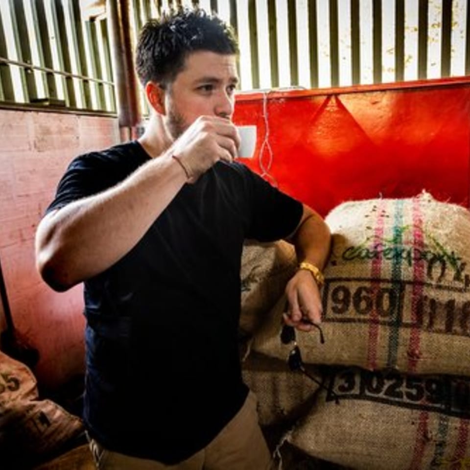 Man in a black shirt sipping coffee from a small white cup, leaning on burlap sacks.