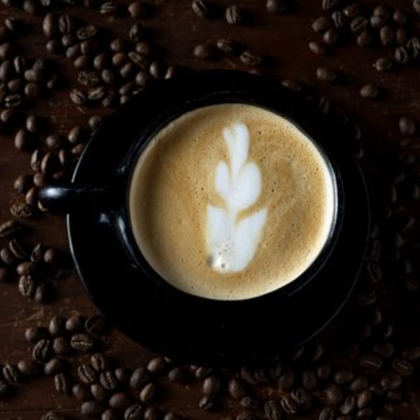 Overhead view of a latte with leaf art in a black cup, surrounded by coffee beans.