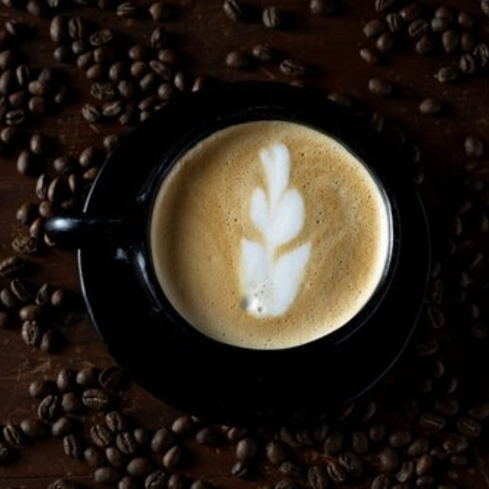 Overhead view of a latte with leaf art in a black cup, surrounded by coffee beans.