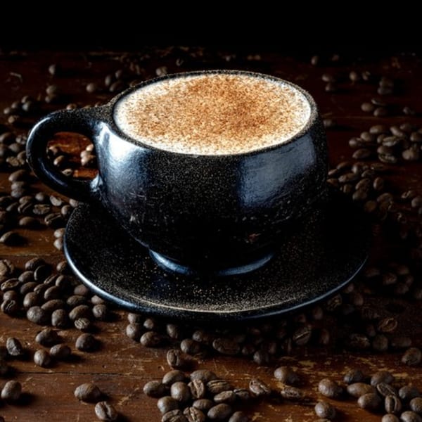 A dark coffee cup with a frothy drink topped with cinnamon, surrounded by coffee beans.