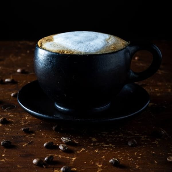 A black ceramic cup of Bolivar Coffee with frothy foam sits on a wooden table with scattered coffee beans.