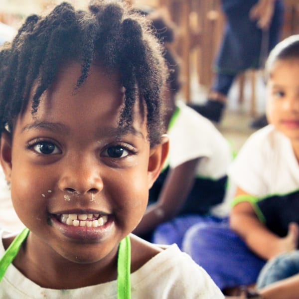 A young child with dark skin and dreadlocks smiles at the camera, with flour on their face.