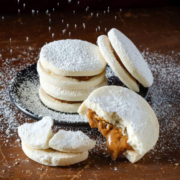 Powdered sugar falling on a stack of alfajores, with one broken open revealing dulce de leche.