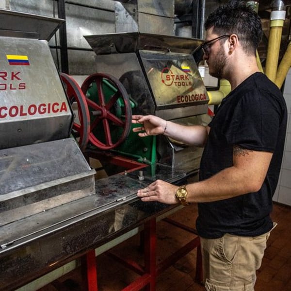 Man in glasses and black shirt operating coffee processing machinery with red wheels.