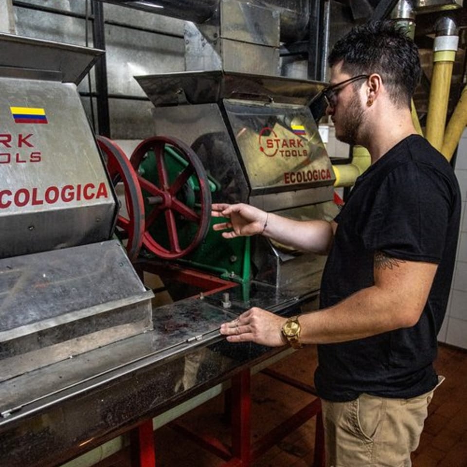 A man in glasses and a black shirt operates coffee processing machinery with Colombian flags.