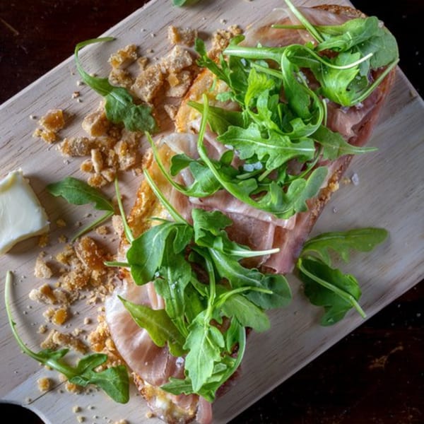 A close-up of a Bolivar Coffee bruschetta with prosciutto, arugula, and crumbled cheese on a wooden board.